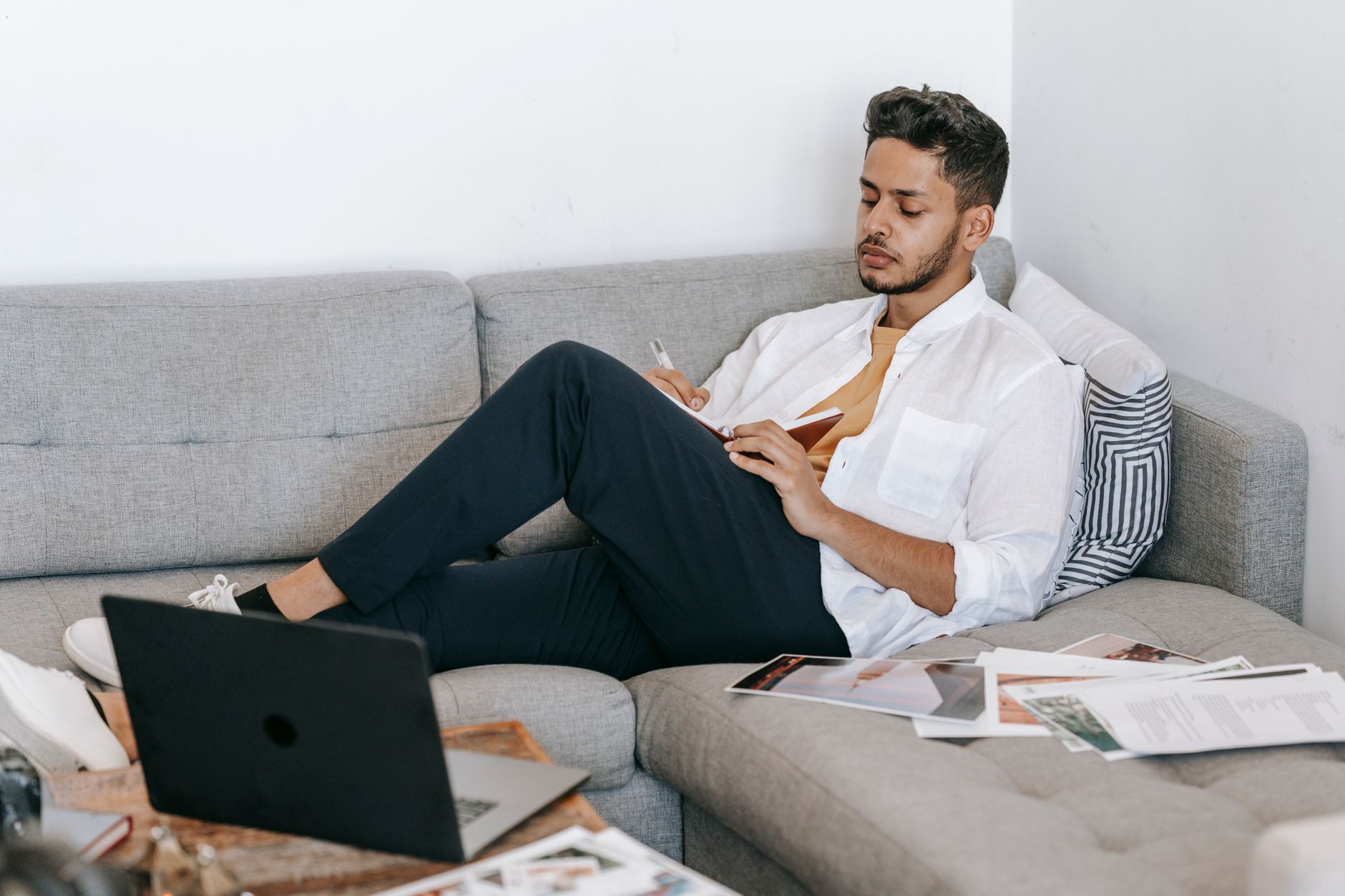 Side view of young ethnic male journalist sitting on sofa and writing ideas for new article in notebook while working with laptop and papers at home