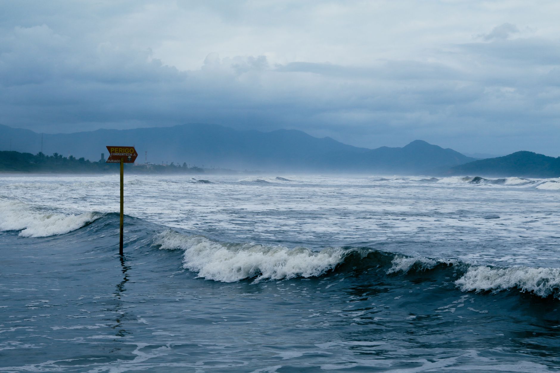 Cloudy coastal scene with warning sign and waves crashing against the shore, conveying sea caution.