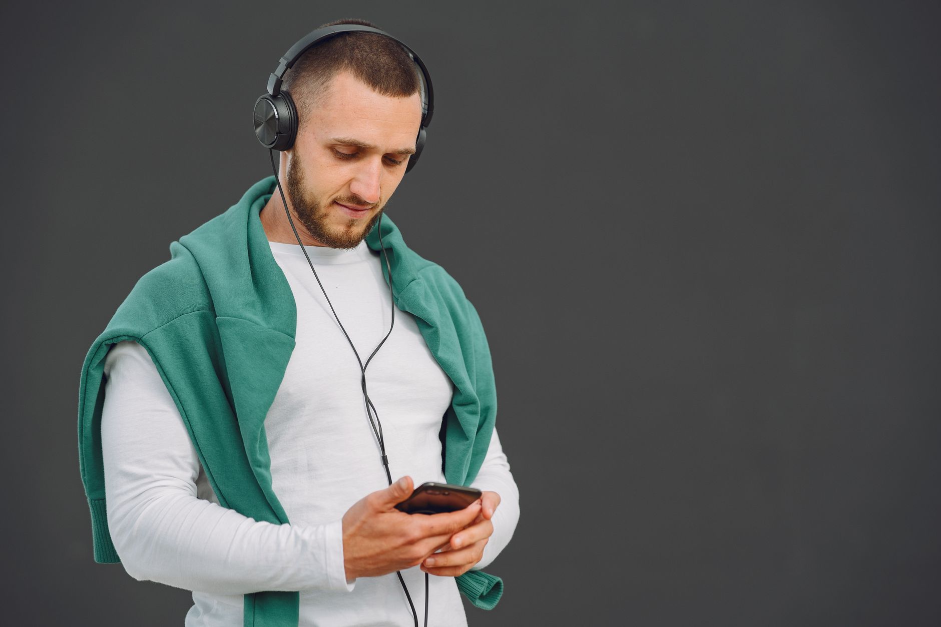 Casual man with headphones listening to music on smartphone against gray background.