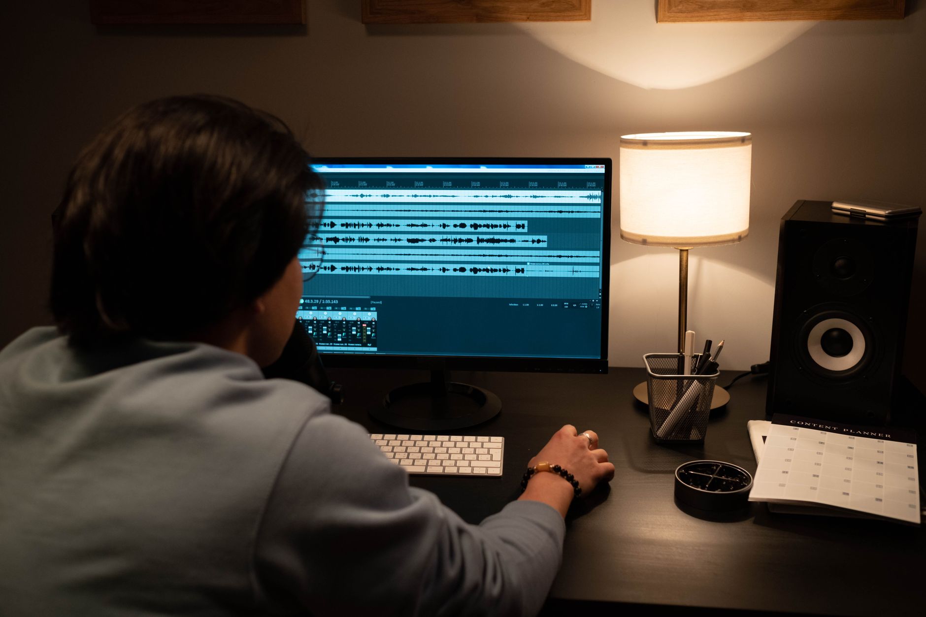 Man wearing a blue sweater editing audio on a monitor in a dimly lit room with a desk lamp.